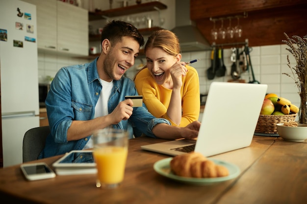 Pareja alegre usando tarjeta de crédito y comprando en línea en casa