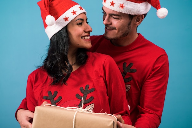 Pareja alegre vistiendo pijamas y sombreros de Navidad miran, sonriendo y sosteniendo un regalo de Navidad