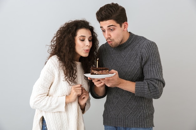 Pareja alegre vistiendo suéteres que se encuentran aisladas sobre la pared gris, celebrando un cumpleaños, sosteniendo un trozo de tarta con vela