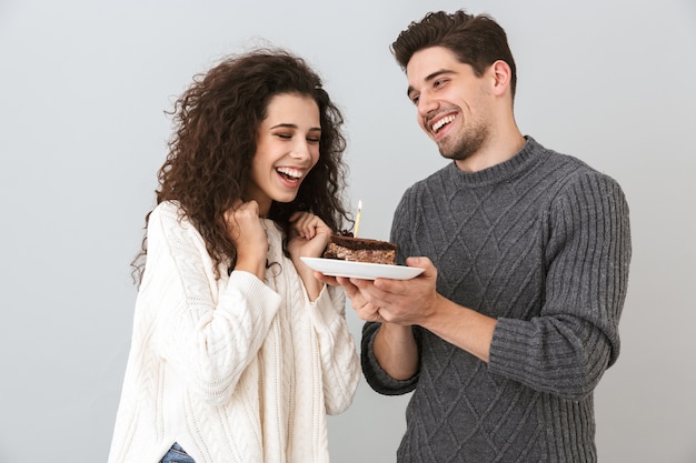 Pareja alegre vistiendo suéteres que se encuentran aisladas sobre la pared gris, celebrando un cumpleaños, sosteniendo un trozo de tarta con vela