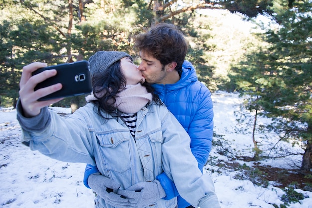 Pareja de amantes disfrutando en la nieve haciendo una selfie mientras se besan.