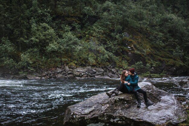 Una pareja de amantes con impermeables verdes, sentados en una roca, en el contexto de una cascada