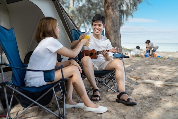 Pareja de amor asiático acampando en la playa de verano. Hombre tocando Ukuleleand junto con una mujer sentada fuera de la tienda.