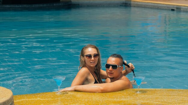 La pareja amorosa se abraza y besa bebiendo licor de alcohol de cóctel azul en la piscina en el hotel al aire libre Retrato de hombre y mujer caucásicos Peinados creativos culturista traje de baño gafas de sol
