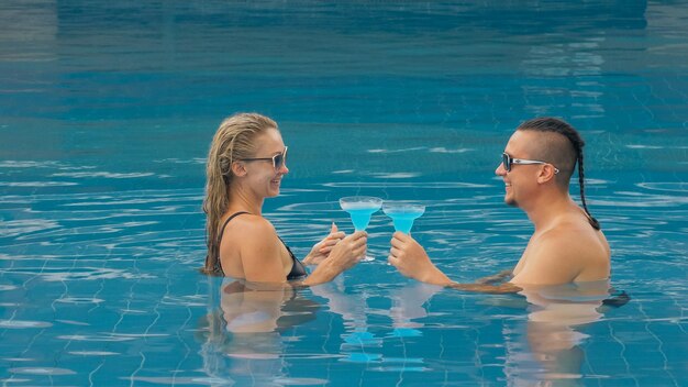 La pareja amorosa se abraza y besa bebiendo licor de alcohol de cóctel azul en la piscina en el hotel al aire libre Retrato de hombre y mujer caucásicos Peinados creativos culturista traje de baño gafas de sol