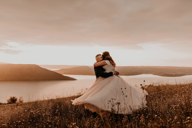 Pareja amorosa boda recién casados en vestido blanco y traje abrazo besos torbellino sobre hierba alta en campo de verano en la montaña sobre el río