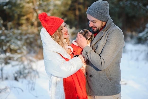 Una pareja amorosa camina de la mano en el fondo de un bosque nevado de invierno Un hombre sostiene un perro Una niña con una chaqueta blanca y un sombrero rojo