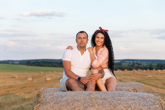 Pareja amorosa en un campo sobre rollos de paja. Joven y mujer divirtiéndose, abrazándose y besándose al atardecer.