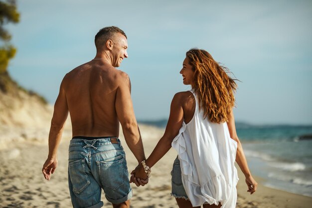 Una pareja amorosa se divierte y camina por la playa de arena vacía. Se miran y sonríen felizmente.