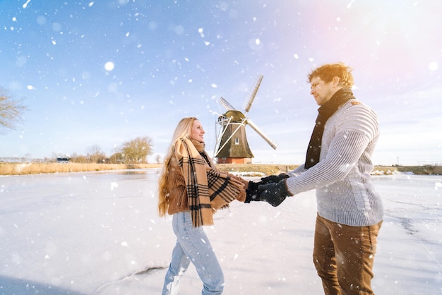 Pareja amorosa divirtiéndose en el hielo en el típico paisaje holandés con molino de viento mujer y hombre patinaje sobre hielo