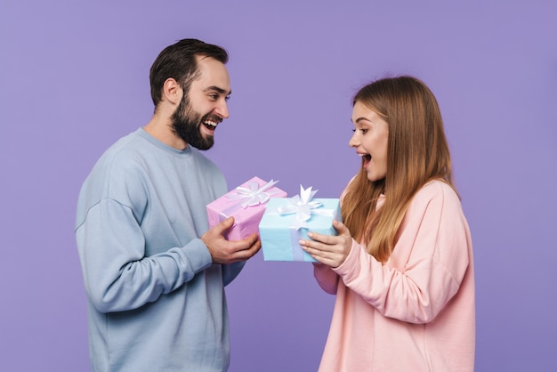 La pareja amorosa joven y positiva emocionada aislada sobre la pared púrpura se da un regalo el uno al otro.