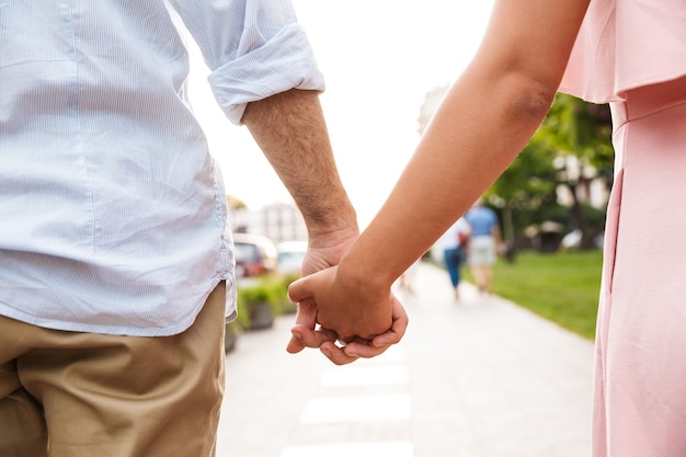 Pareja amorosa joven recortada caminando por la calle al aire libre cogidos de la mano.