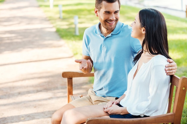 Pareja amorosa en el parque. Vista lateral de la alegre joven pareja amorosa sentados en el banco juntos y hablando