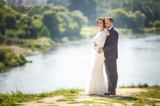 Una pareja amorosa de recién casados camina cerca de un río ancho en un día soleado de verano