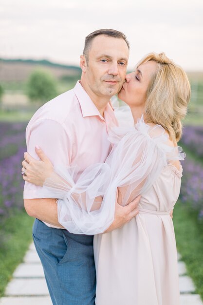 Pareja amorosa romántica madura caminando en el campo de lavanda. Pareja de mediana edad caucásica feliz soñadora en la naturaleza, de pie cogidos de la mano. Mujer besando a su hombre. Disfrutando el momento.