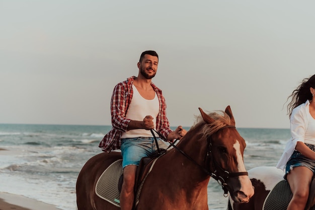 Una pareja amorosa con ropa de verano montando a caballo en una playa de arena al atardecer. Mar y atardecer de fondo. Enfoque selectivo. foto de alta calidad