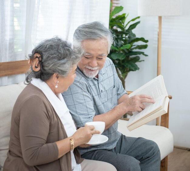 Pareja de ancianos asiáticos mayores sentados en el sofá leyendo el libro juntos en casa