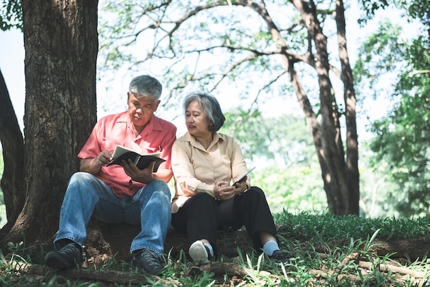 Pareja de ancianos asiáticos sentados bajo un árbol en el parque y mirando leyendo un libro diario