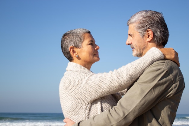 Una pareja de ancianos canosos abrazándose a la orilla del mar en un soleado día de otoño. Señora de mediana edad de pelo corto y anciano barbudo mirándose el uno al otro. Vista lateral. Relación, amor, concepto de viaje.