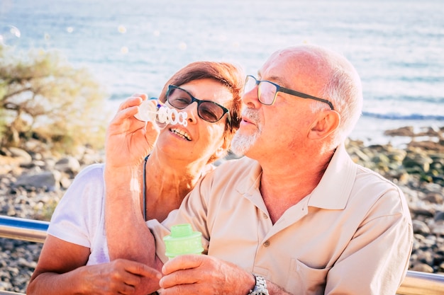 Pareja de ancianos casados sentarse en la playa soplando una burbuja de jabón con el mar de fondo