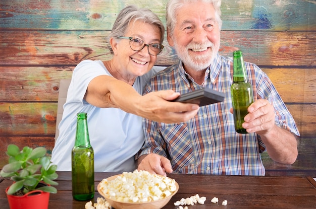 Pareja de ancianos divirtiéndose viendo un partido de fútbol en la televisión. Botellas de cerveza y palomitas de maíz en la mesa