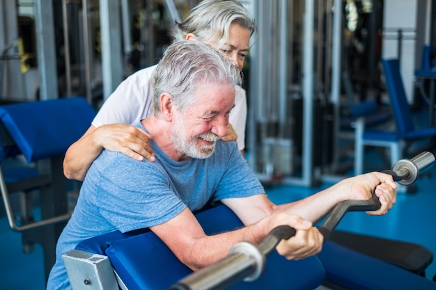 Pareja de ancianos en el gimnasio haciendo ejercicio juntos - mujer madura mirando y ayudando a su marido sosteniendo un barr y levantando pesas