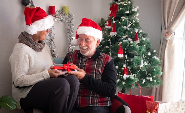 Una pareja de ancianos con gorro de Papá Noel sentados juntos en el mismo sillón. Sosteniendo una tableta recibida como regalo de Navidad. Regalos y árbol de Navidad en el fondo