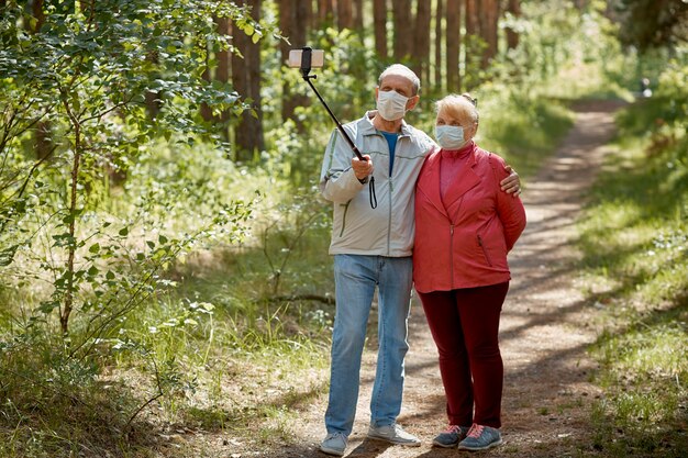 Una pareja de ancianos con máscaras protectoras se toma una selfie en un teléfono inteligente y camina por el parque
