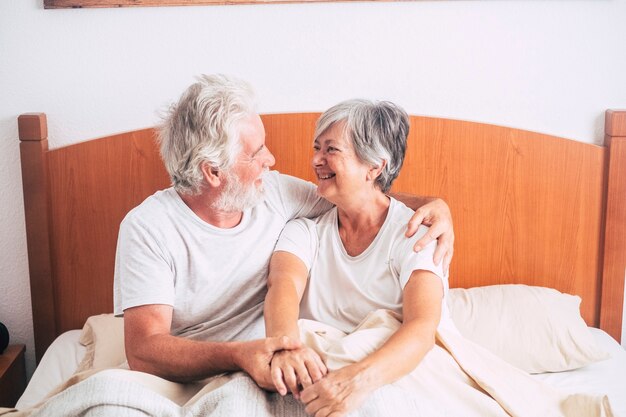 Pareja de ancianos mirando y sonriendo con amor y afecto mientras ella lo besa - adultos jubilados y maduros casados en el dormitorio por la mañana antes de levantarse