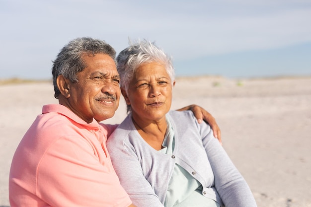 Una pareja de ancianos multirracial pensativa mirando hacia otro lado mientras se relaja en la playa en un día soleado