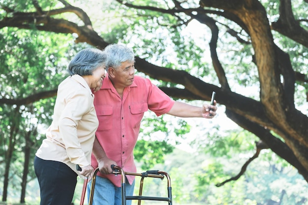 Pareja de ancianos de pie con un bastón descansando en el jardín y usando el teléfono móvil para tomar una foto