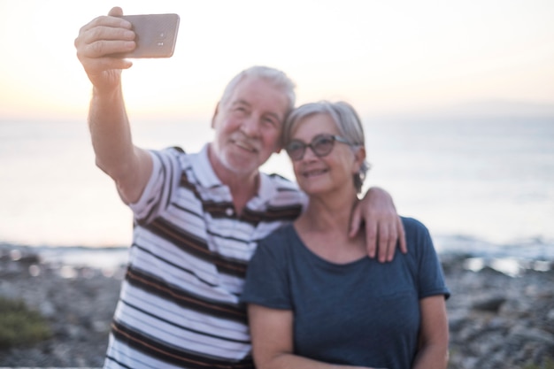 Pareja de ancianos en la playa tomando una foto juntos - mujer con gafas y hombre jubilado - selfie en la playa - divertirse y disfrutar - caucásico