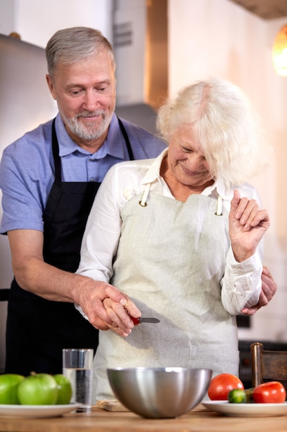 Pareja de ancianos preparando ensalada de verduras en la cocina, el hombre guapo de pelo gris ayuda a la esposa a cocinar, va a tomar un desayuno saludable