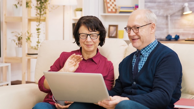 Pareja de ancianos sentados en el sofá sosteniendo el portátil durante una videollamada. Pareja saludando a la computadora portátil.