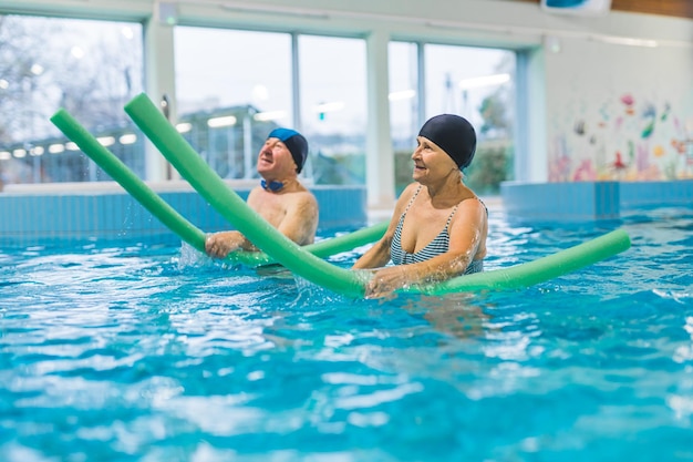 Una pareja de ancianos sonrientes haciendo ejercicio en la piscina con fideos de natación personas jubiladas felices juegan en