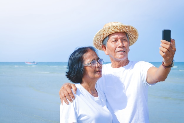 Una pareja de ancianos usa una camisa blanca para ir al mar. De pie sosteniendo el teléfono para tomar fotos juntos