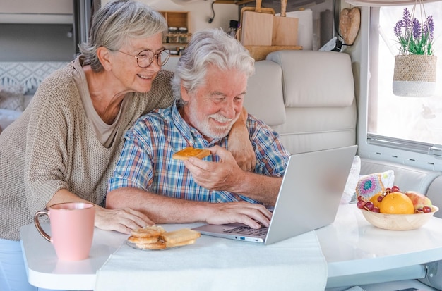 Pareja de ancianos en vacaciones de viaje dentro de una autocaravana disfrutando del desayuno juntos Feliz pareja caucásica de ancianos en la vida de la camioneta mientras navega en la computadora portátil Luz brillante desde la ventana