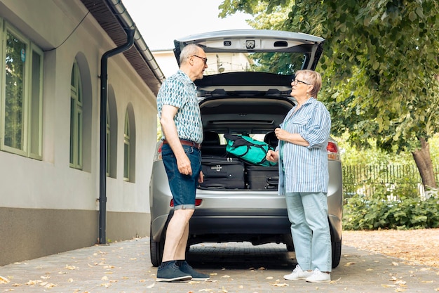 Pareja de ancianos yendo de vacaciones, cargando maleta y equipaje en el maletero del automóvil en casa. Viajar en automóvil con equipaje y bolsas, dejando un viaje de aventura en el paisaje urbano de Onubran.