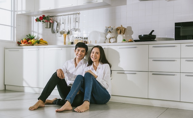 Pareja asiática atractiva joven vestida con camisa blanca y jeans sentados juntos en el piso de la cocina.