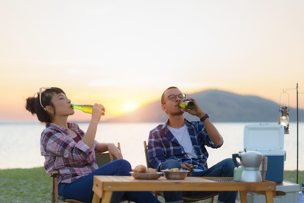 Una pareja asiática bebiendo cerveza de una botella en su área de campamento con el lago en el fondo