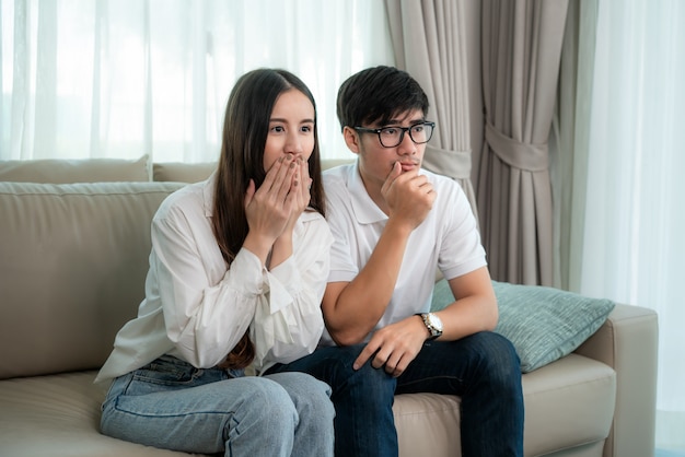 Pareja asiática hombre y mujer mirando y disfrutando de la película de televisión terror sentado en un sofá juntos en la sala de estar en casa. Concepto de relajación y recreación de estilo de vida familiar.