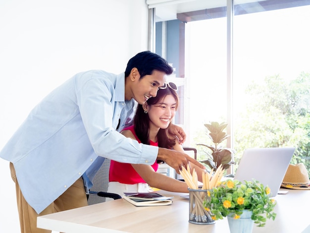 Pareja asiática joven mujer y hombre mirando y señalando la pantalla de la computadora portátil en el escritorio juntos para reservar viajes habitación de hotel e información de viaje en la oficina feliz concepto de vacaciones de verano