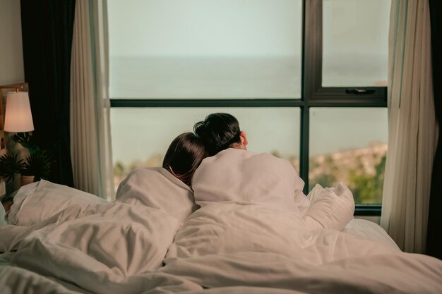 Pareja asiática sentada muy cerca admirando la vista al mar mirando por la ventana del dormitorioPareja en el día de San ValentínAmor de parejaRelación y concepto de pareja