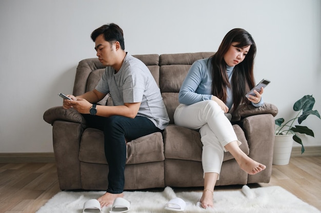 Una pareja asiática usando el teléfono móvil mientras se sientan de espaldas el uno al otro en el sofá