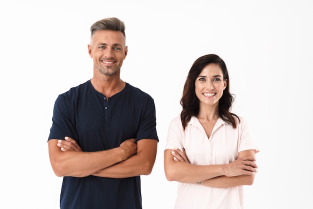 Pareja atractiva alegre vistiendo ropa casual que se encuentran aisladas sobre una pared blanca, con los brazos cruzados