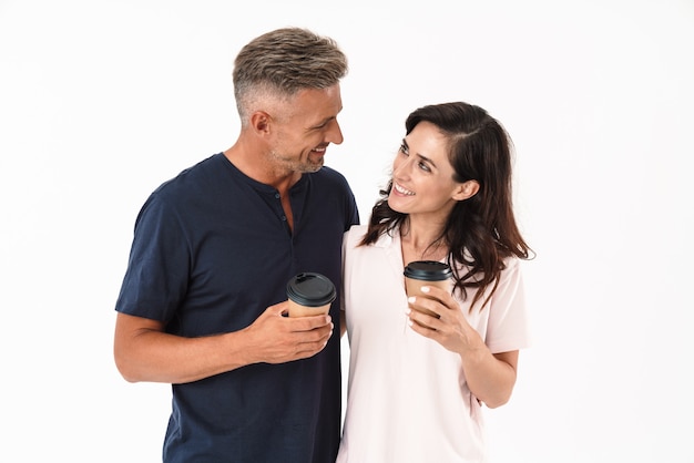 Pareja atractiva alegre vistiendo ropa casual que se encuentran aisladas sobre una pared blanca, sosteniendo tazas de café para llevar, mirando el uno al otro