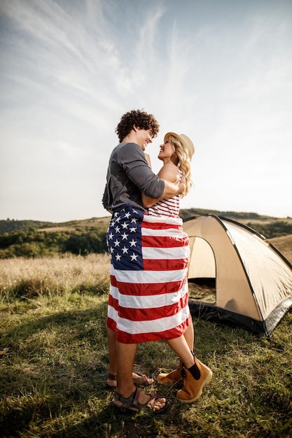 Una pareja atractiva enamorada envuelta en bandera americana disfrutando frente a una carpa en el campamento.