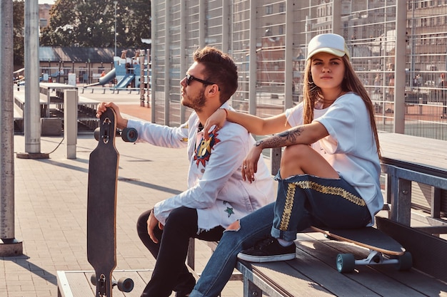 Pareja atractiva de jóvenes hipsters vestidos de moda con patinetas sentados en un banco en el complejo deportivo de la ciudad en un día soleado, con tonos cálidos.