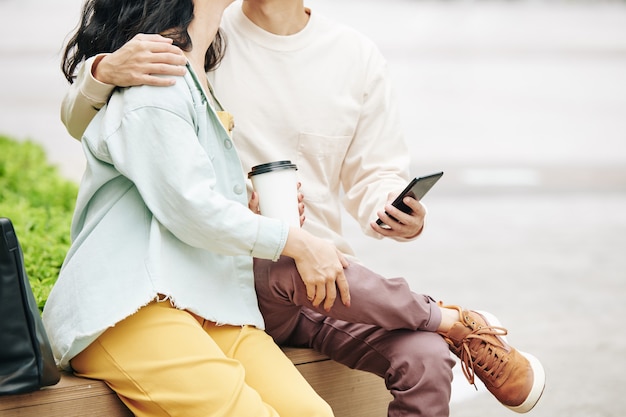 Pareja besándose con teléfono inteligente y tomar café sentado en un banco en el parque y besos