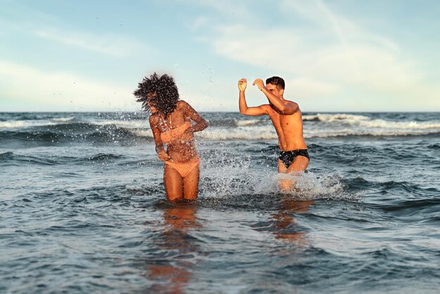 Pareja birracial comprometida de vacaciones jugando juntos salpicando agua bañándose entre las olas jóvenes multiétnicos disfrutando del verano sin preocupaciones